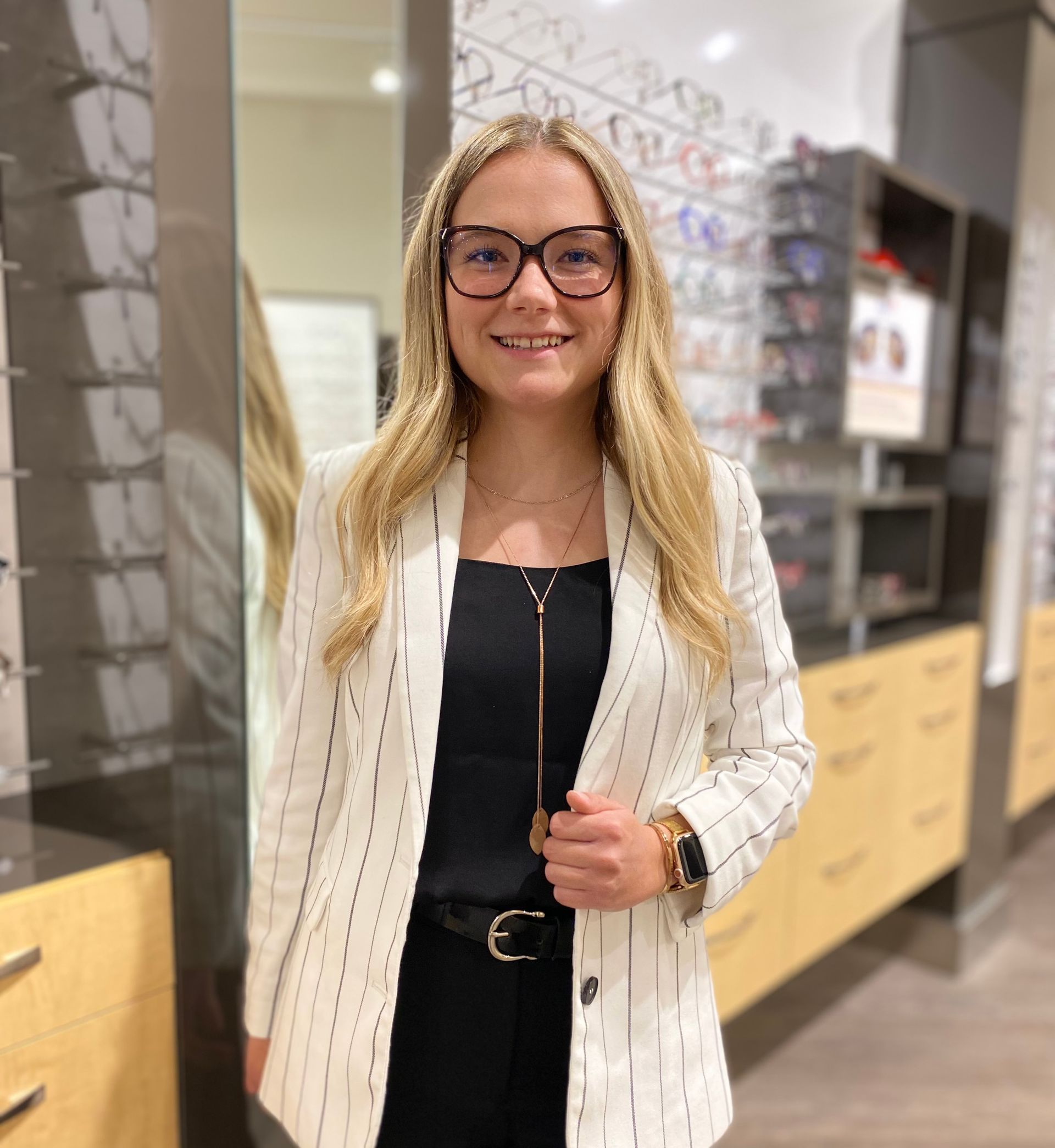 Femme portant des lunettes et un blazer à fines rayures dans un magasin d'optique, souriante.