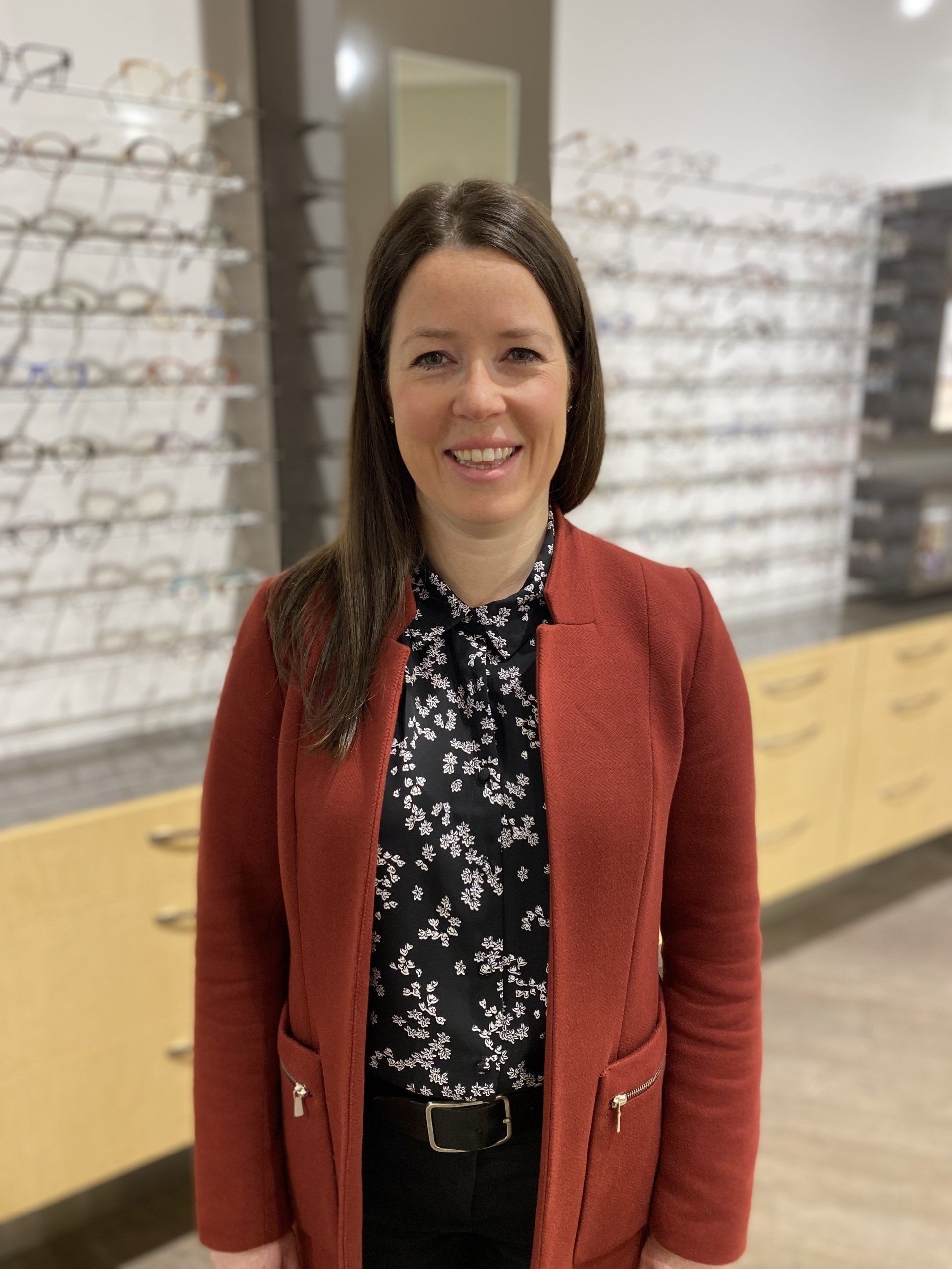 Une femme en veste couleur rouille sourit, debout dans un magasin d'optique. Chemisier à fleurs noires et blanches.