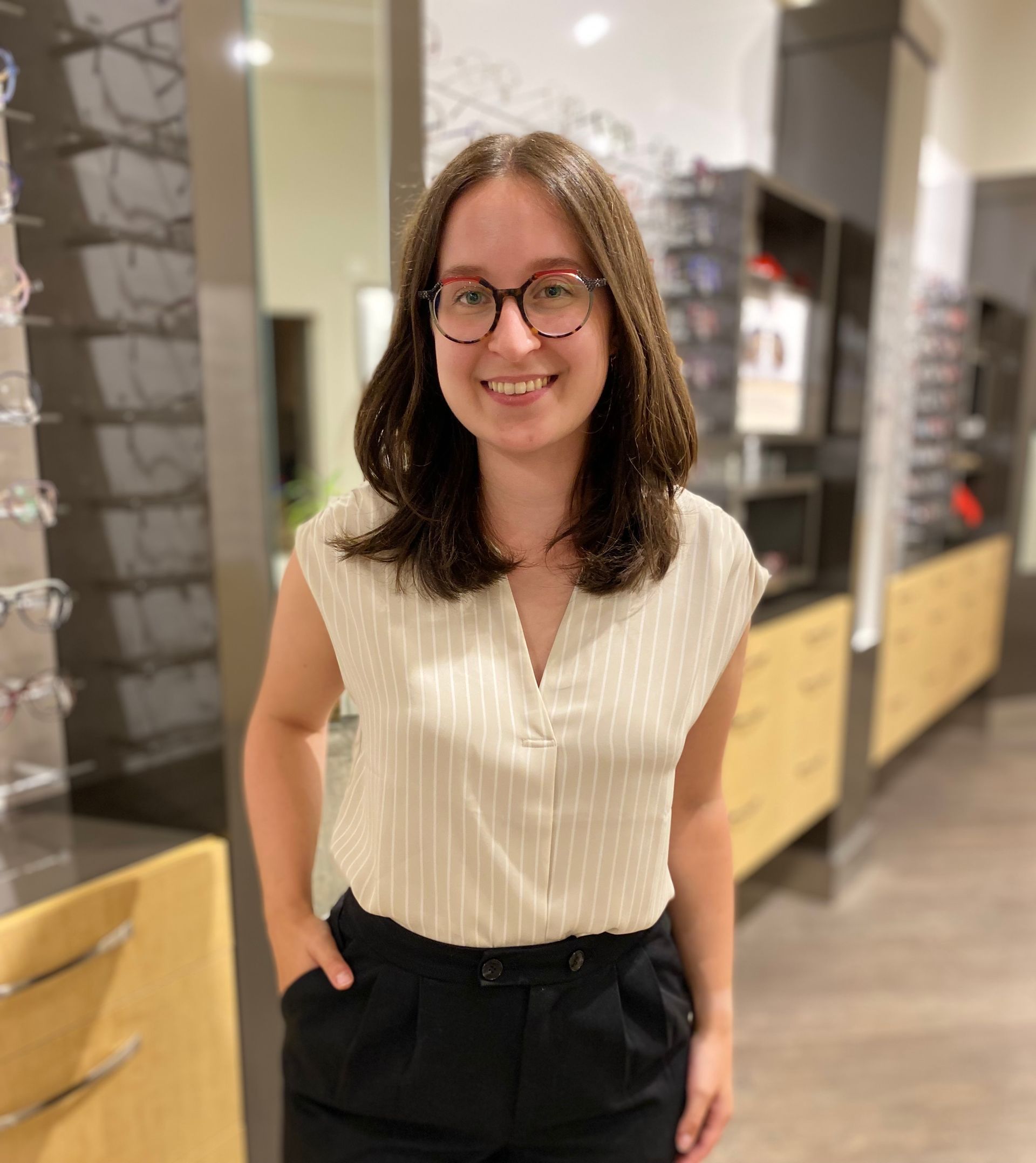 Une femme portant des lunettes sourit, debout dans un magasin d'optique.