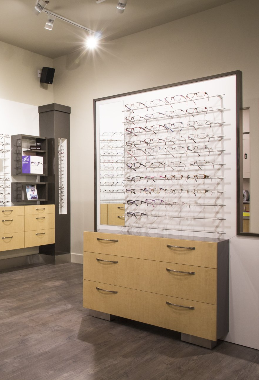 Des montures de lunettes exposées sur un mur dans un magasin, au-dessus de tiroirs en bois clair, d'un socle gris foncé et d'un sol en bois.