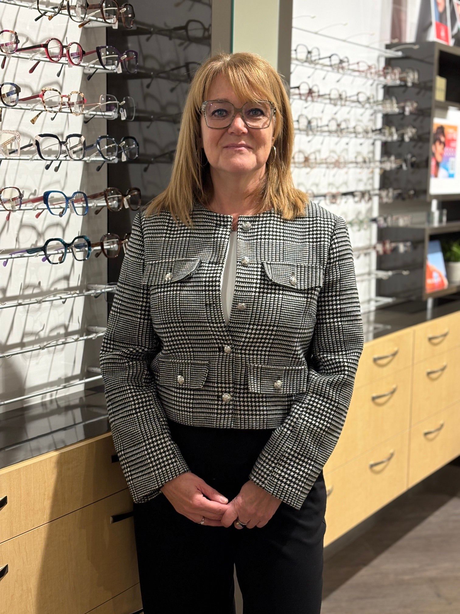 Une femme portant des lunettes et un blazer noir se tient dans un magasin d'optique, souriante.