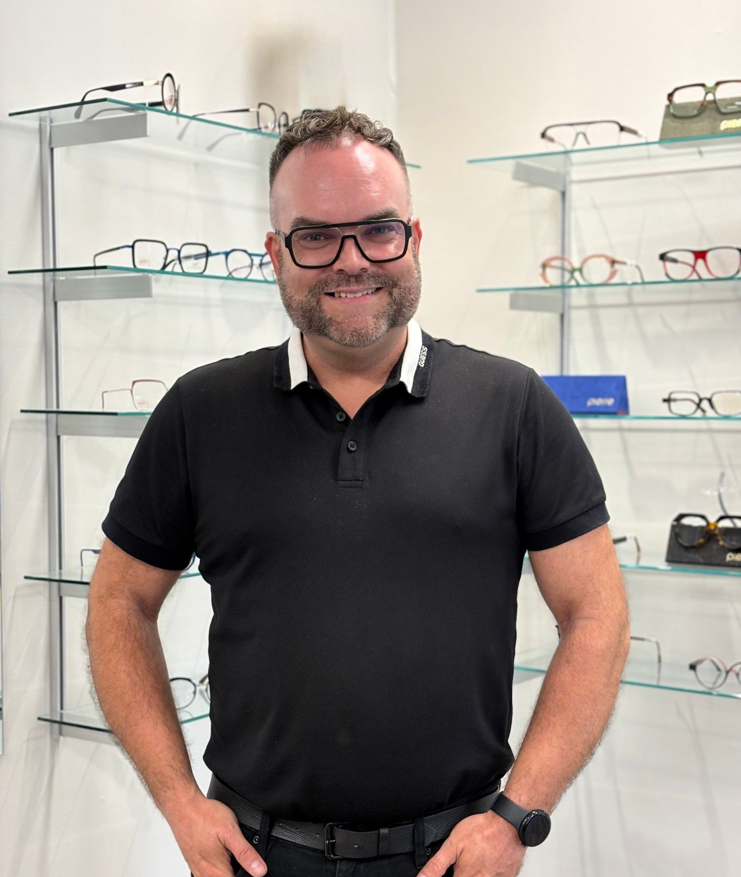 Un homme portant des lunettes sourit, debout dans un magasin d'optique ; des étagères de montures sont visibles en arrière-plan.