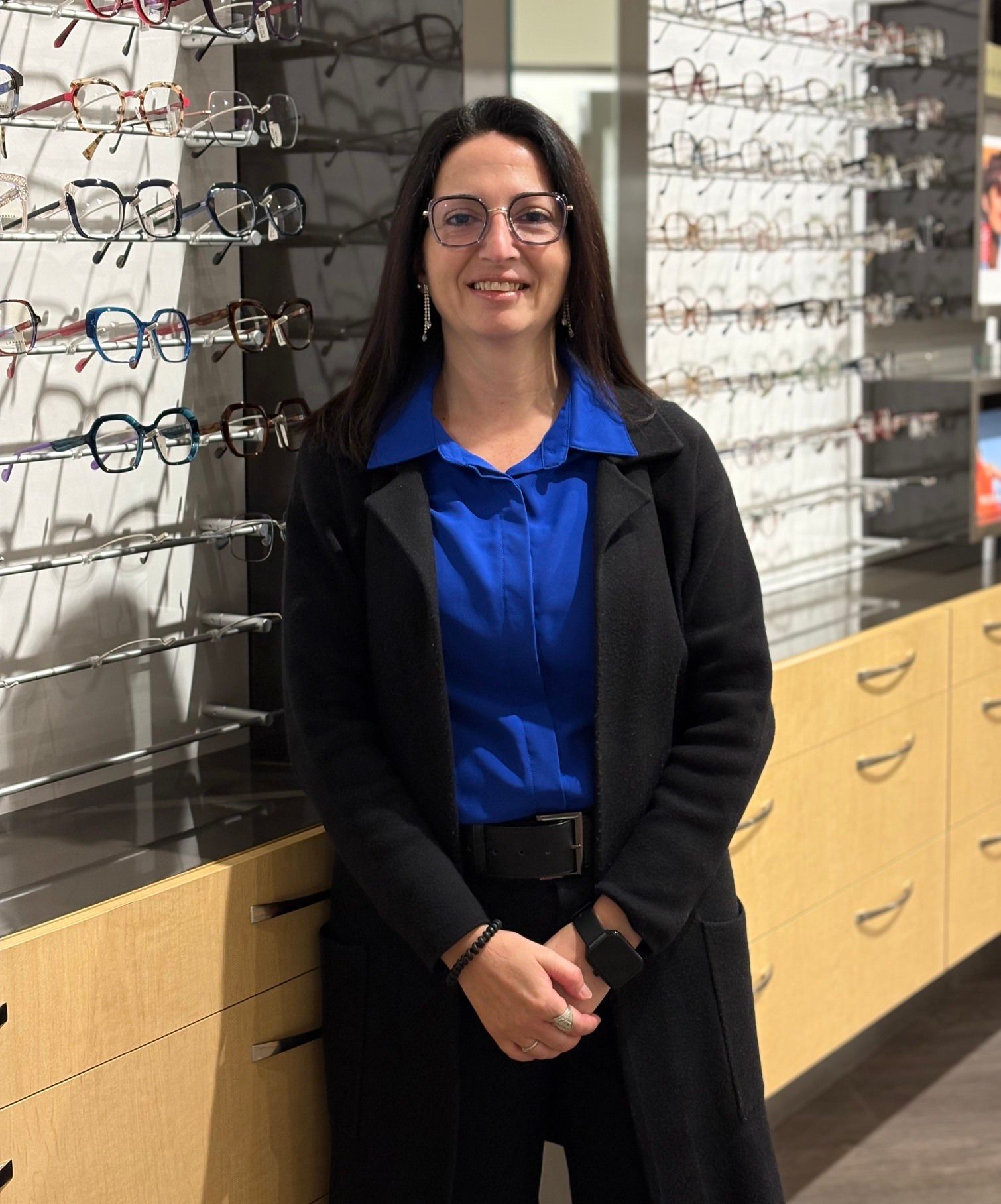 Une femme portant des lunettes se tient dans un magasin d'optique. Elle porte un cardigan noir sur un chemisier rose pâle. Des lunettes sont exposées en arrière-plan.