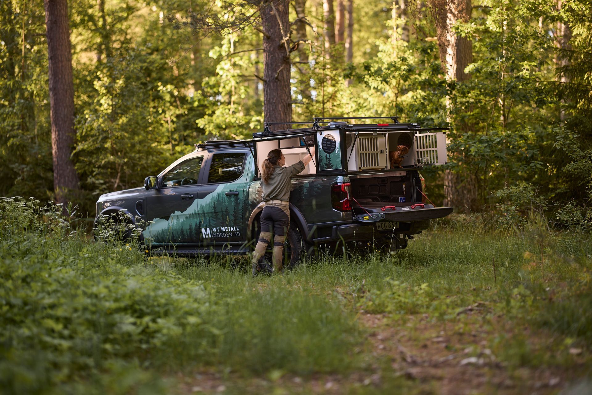 En Labrador är  ute och går i skogen med sin ägare. i Bakgrunden står en pickup.