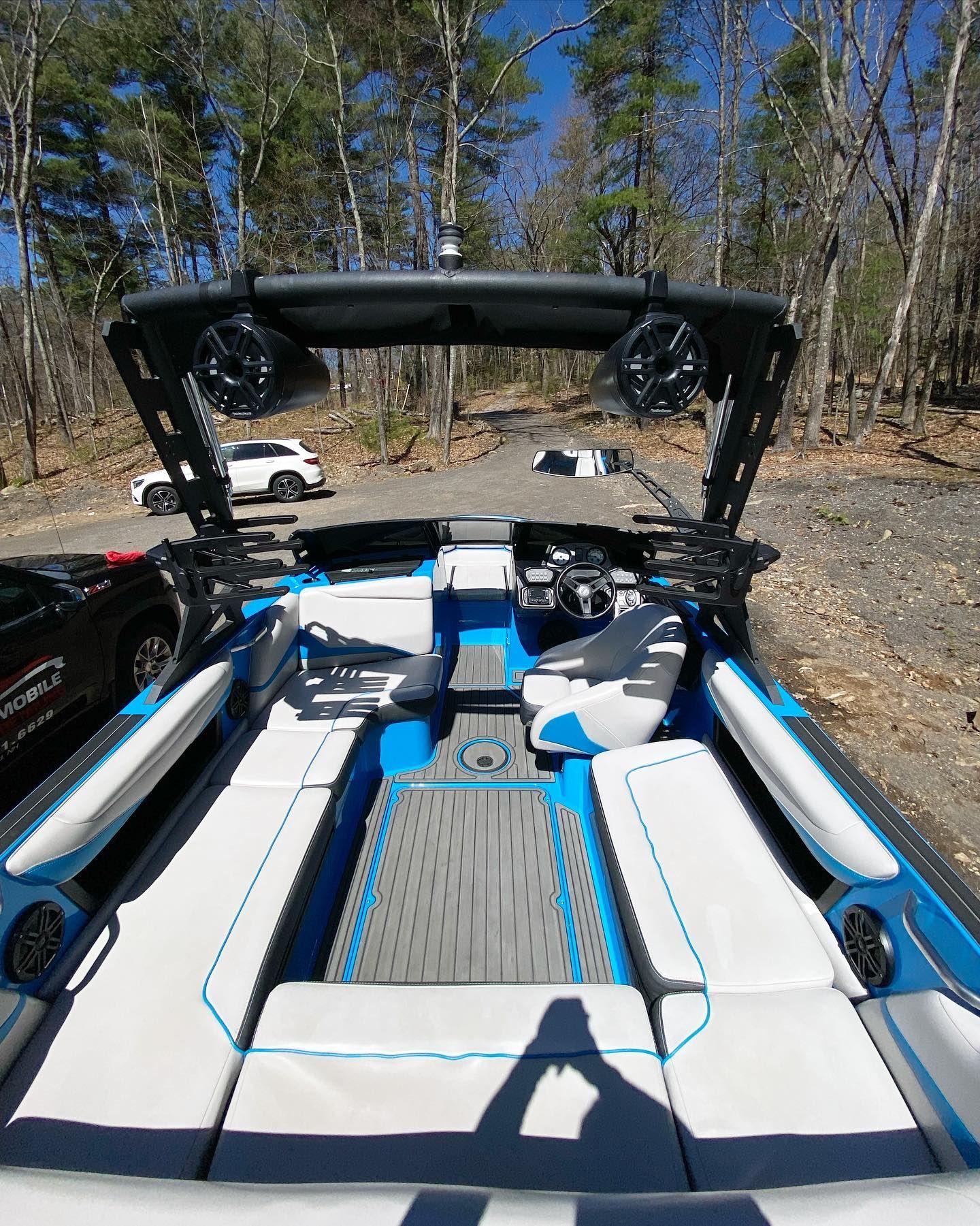 Black-and-white boat cockpit with wakeboard tower, parked outdoors in a wooded lot