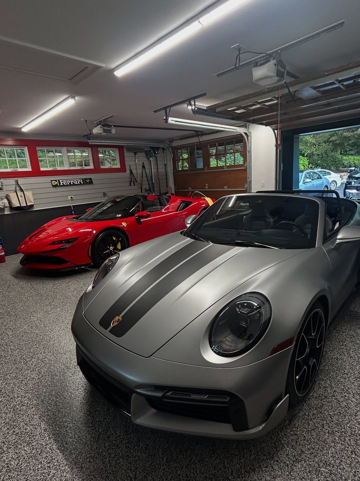 Silver Porsche in a garage with a red sports car behind it, under bright overhead lights.