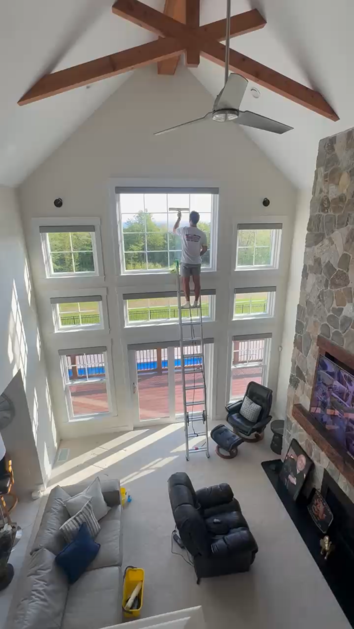 Tall living room with large windows, a ceiling fan, sofa, recliner, and a person standing on a ladder by the window.