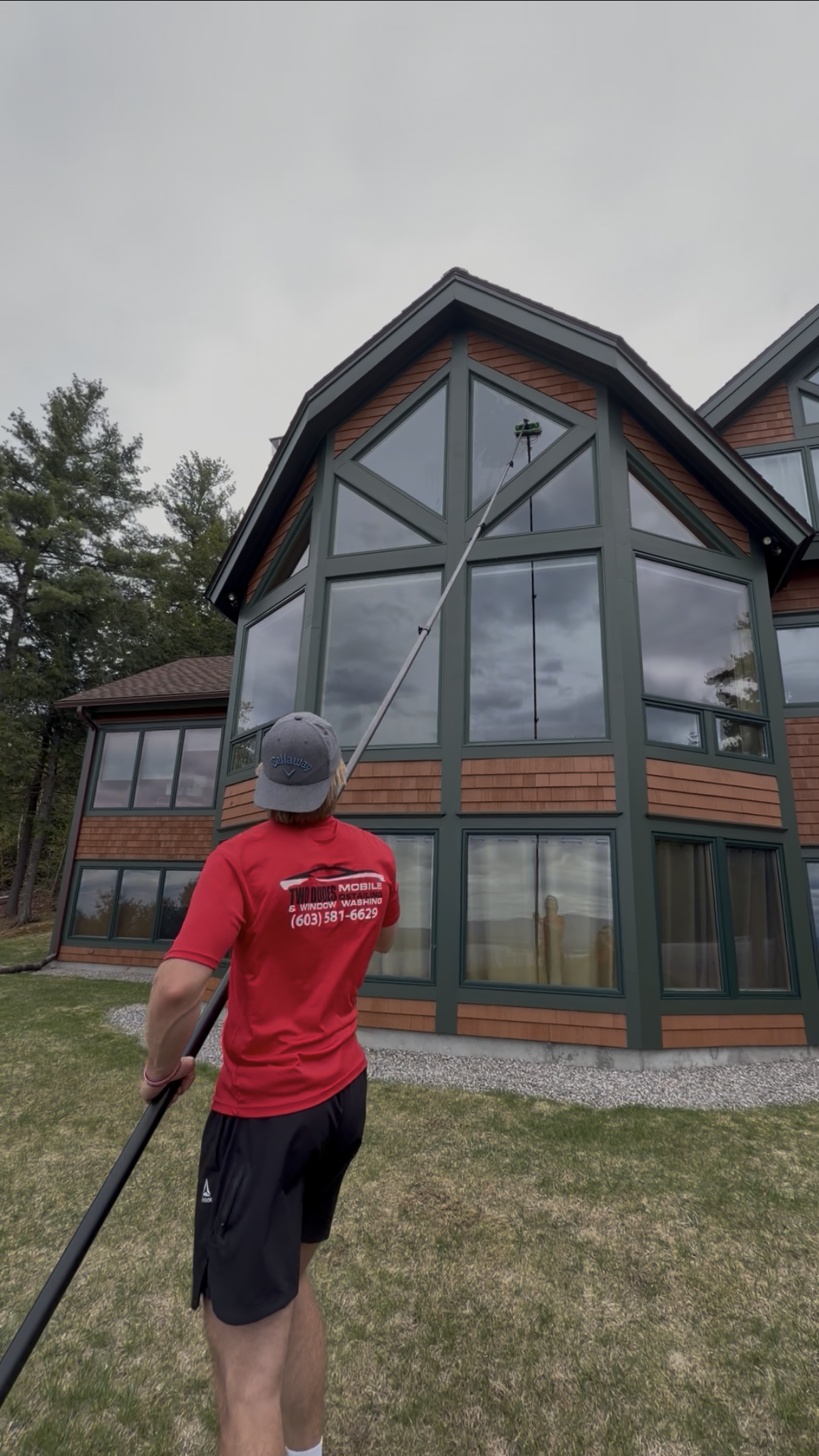 Person in red shirt and gray cap standing on grass beside a modern house with large windows and steep roof.