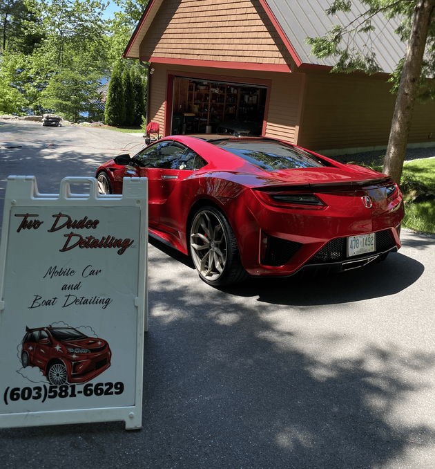 Red sports car parked beside a roadside sign outside a building in a shady driveway