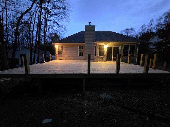 A new wooden deck attached to a house with lights on. Dusk setting with trees in the background.
