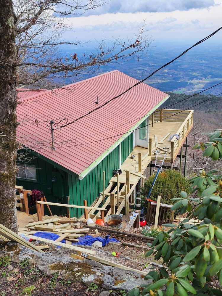 Green cabin with red roof and wooden deck on a mountain. Construction materials scattered. Cloudy sky, valley view.