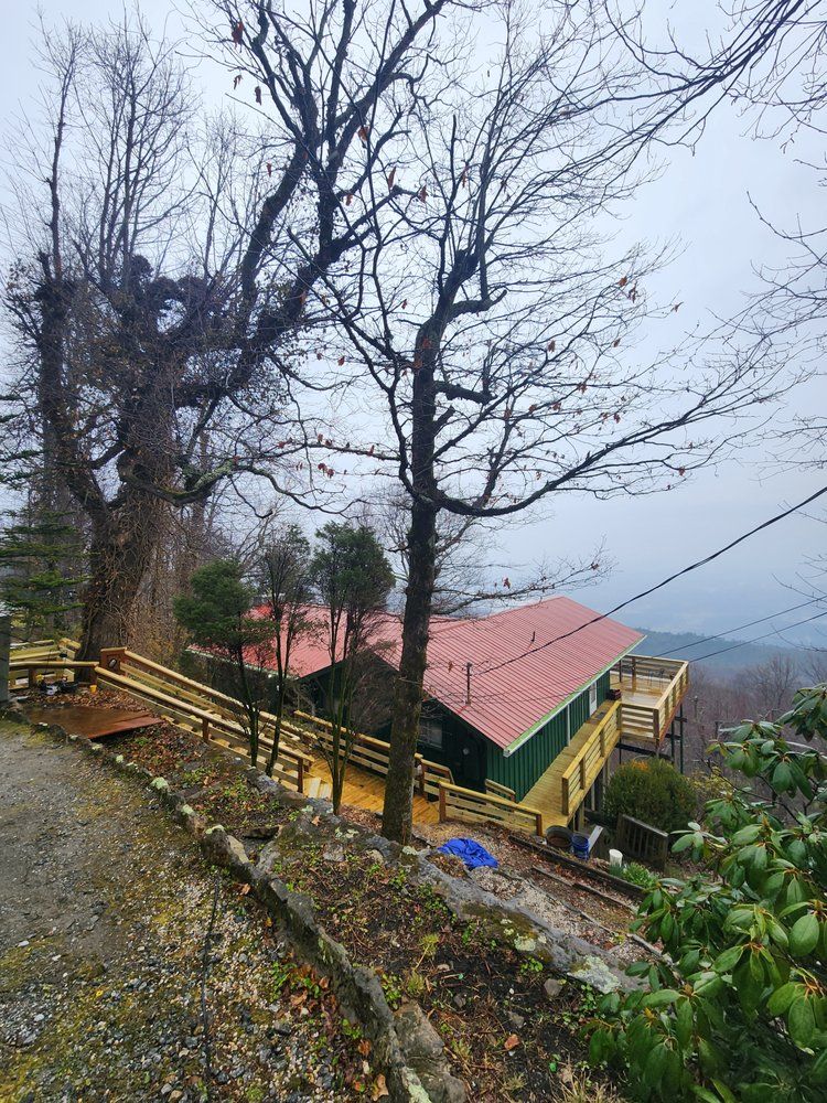 Cabin on a hillside with a red roof, surrounded by trees and a cloudy sky.