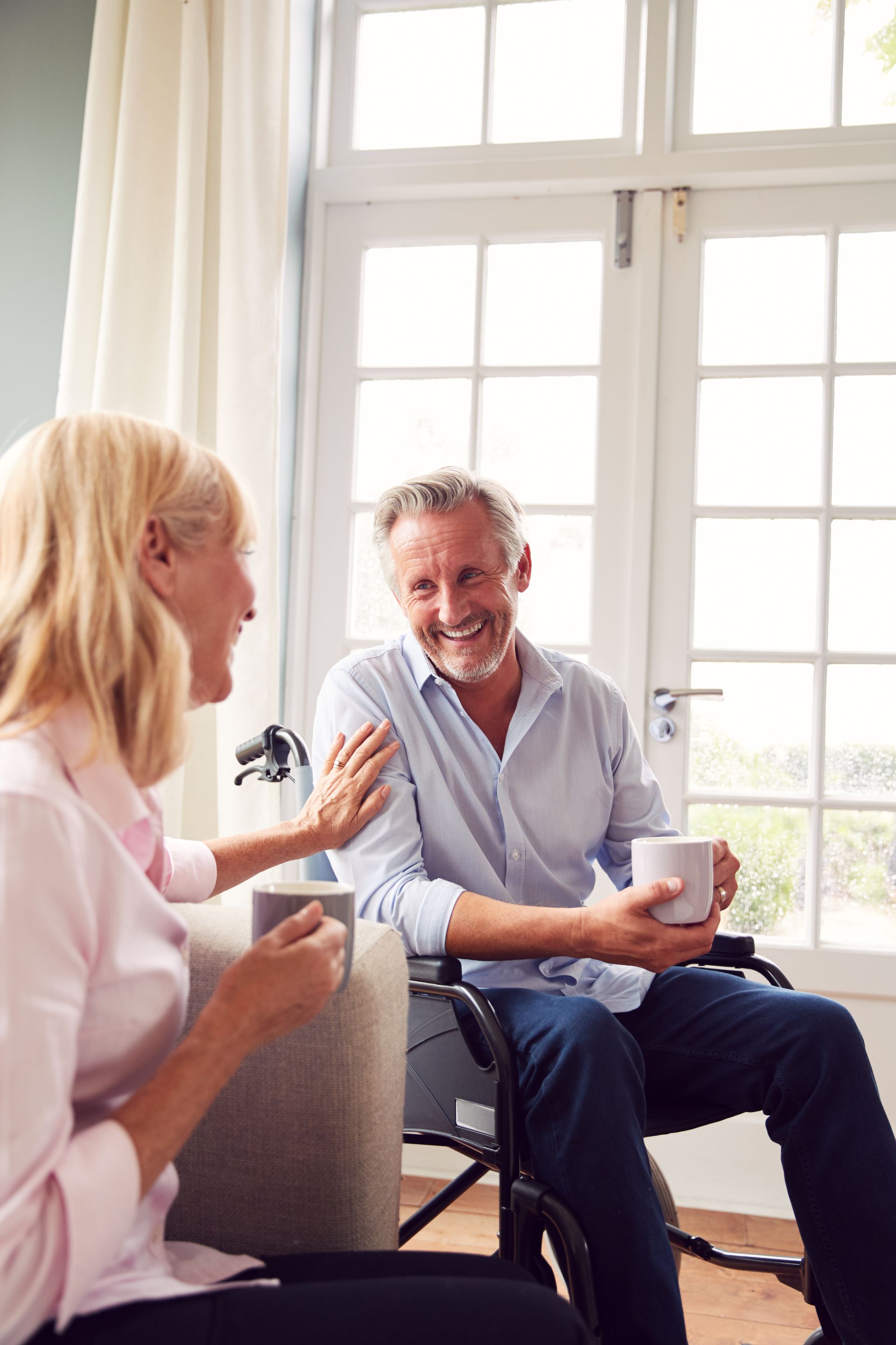 Woman and man smiling, talking, holding mugs. Man in wheelchair, by window.