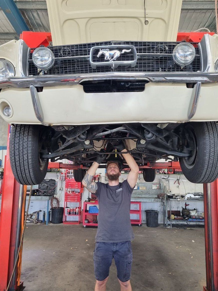 A Man Is Working Under A Mustang On A Lift In A Garage — Dancers Car Care In Ballina, NSW