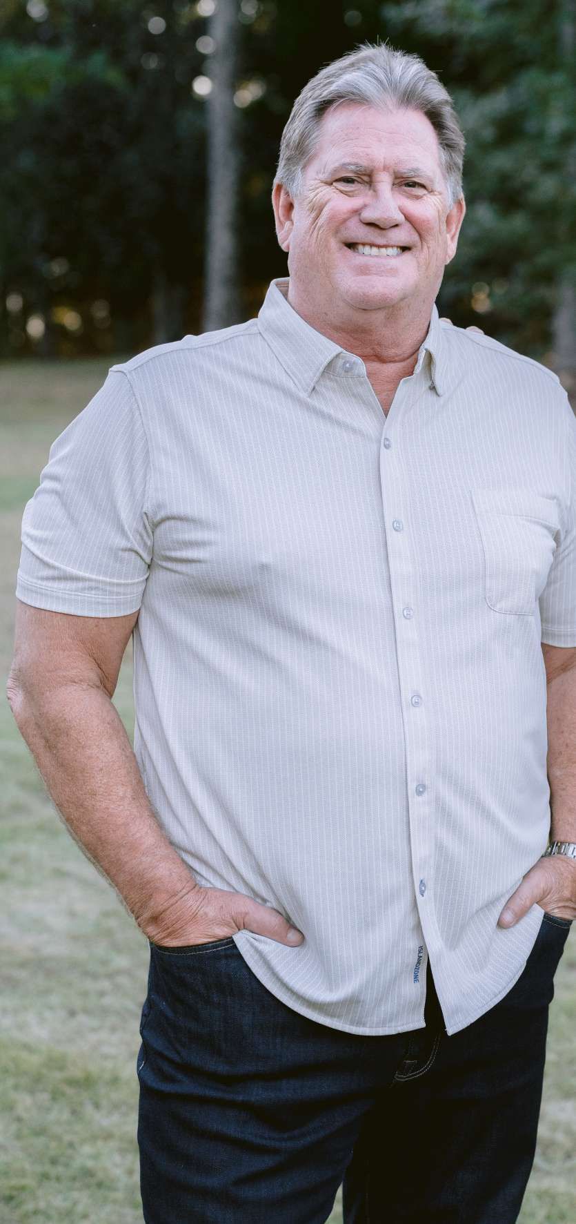 A smiling person with short hair, wearing a light-colored, button-down shirt and dark pants, standing in a park.