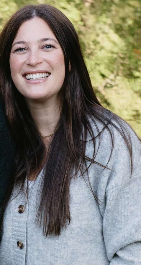 A smiling person with long dark hair, wearing a gray button-down cardigan, poses outdoors against a blurred green background.