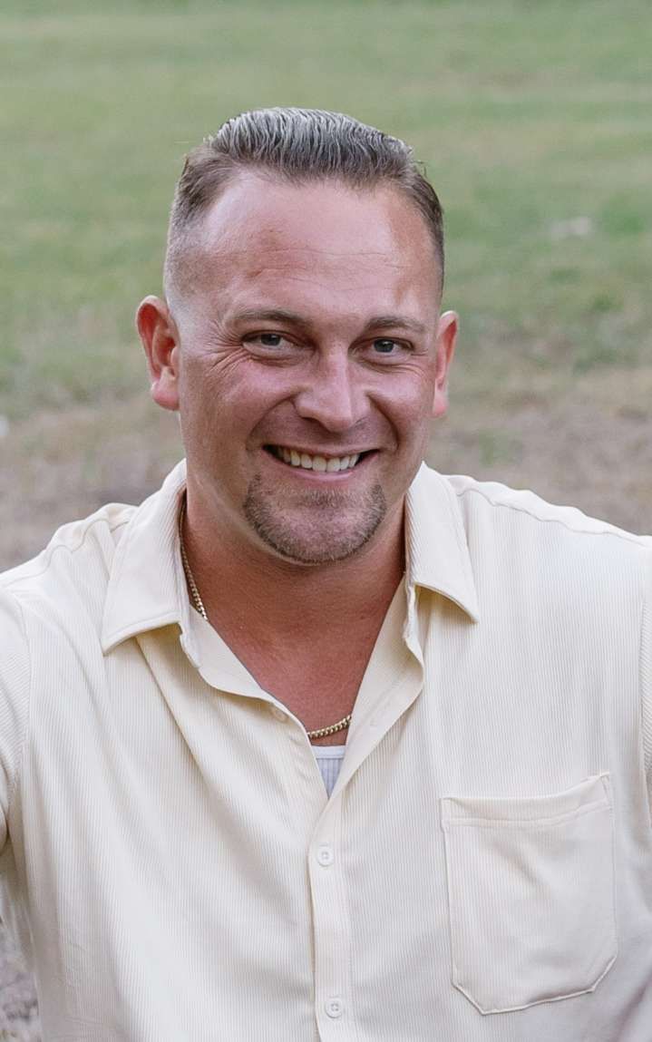 A person with a short, faded haircut and a light goatee smiles at the camera, wearing a light yellow button-down shirt.