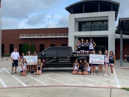 A group of cheerleaders in matching uniforms poses with a pickup truck in front of North Forsyth High School.