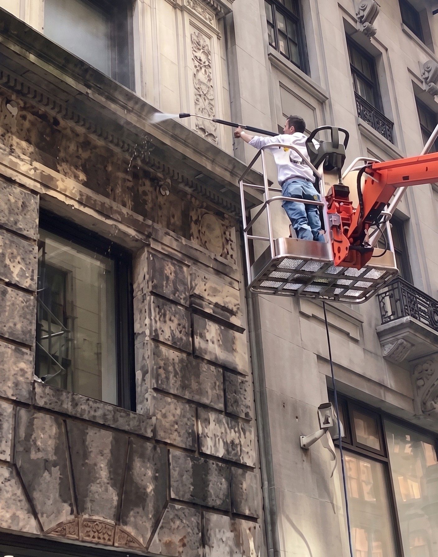 Worker in a lift basket washing a tall stone building facade with a pressure washer.