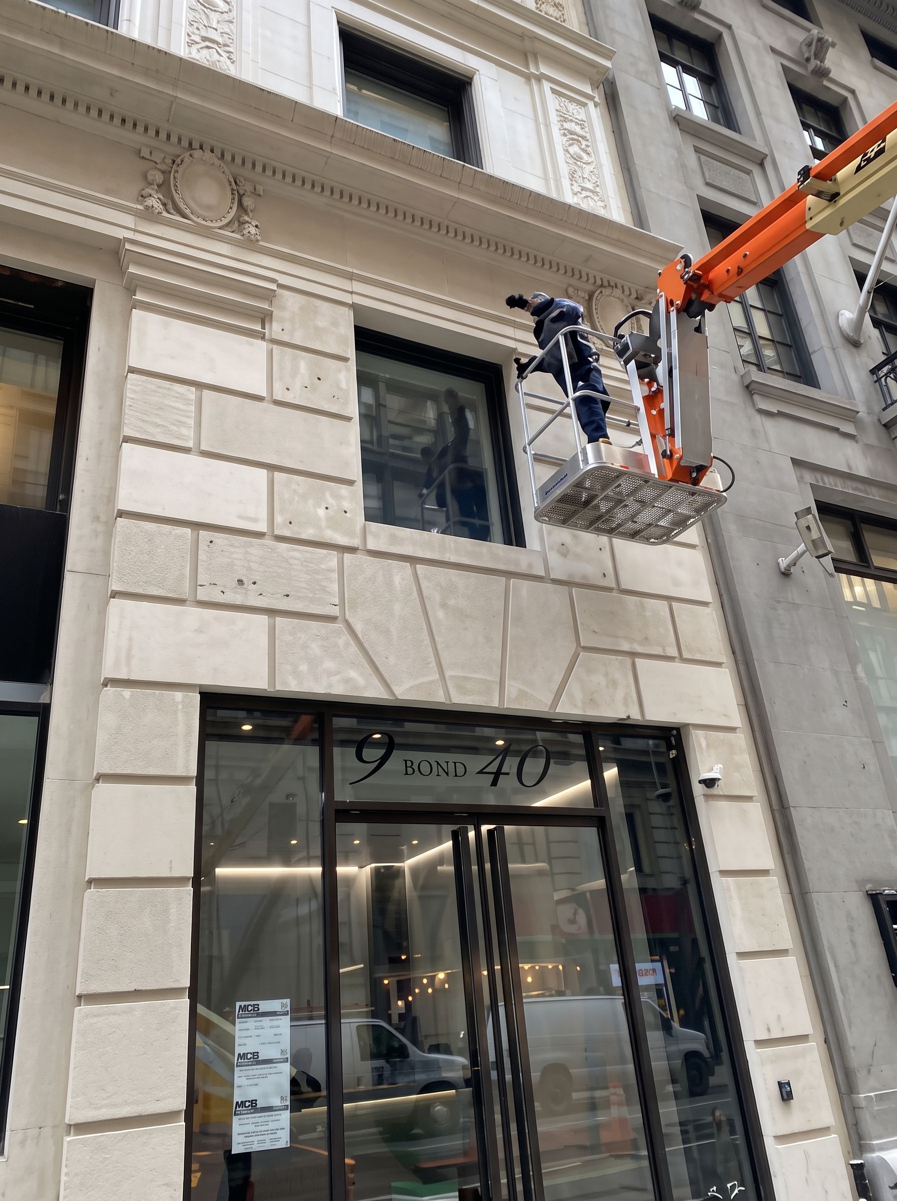 Worker in a cherry picker cleaning a stone building facade above a storefront.