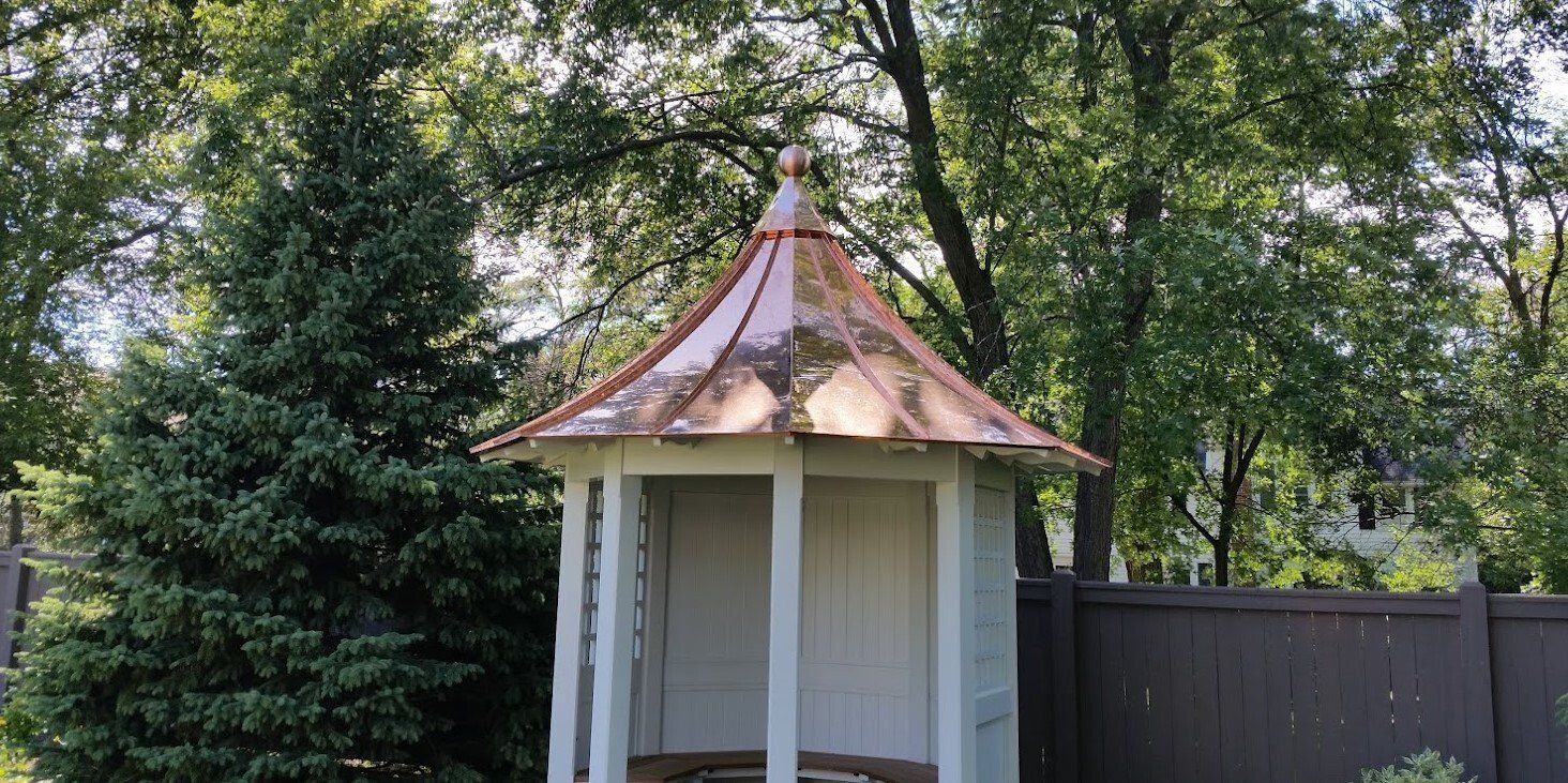 Small white gazebo with a rusted copper roof in a leafy backyard