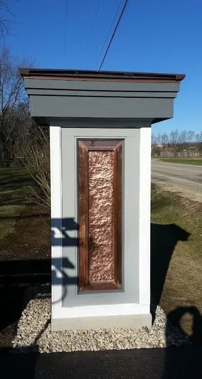 A copper-paneled pillar with a gray top and base, set in gravel next to a road, under a bright sky. Small white utility kiosk with a copper panel door and dark roof beside a gravel path.