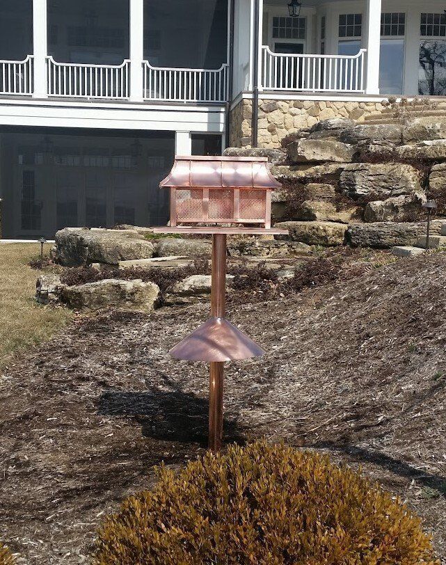 Rusty metal bird feeder in a landscaped yard with stone retaining wall and house porch behind it