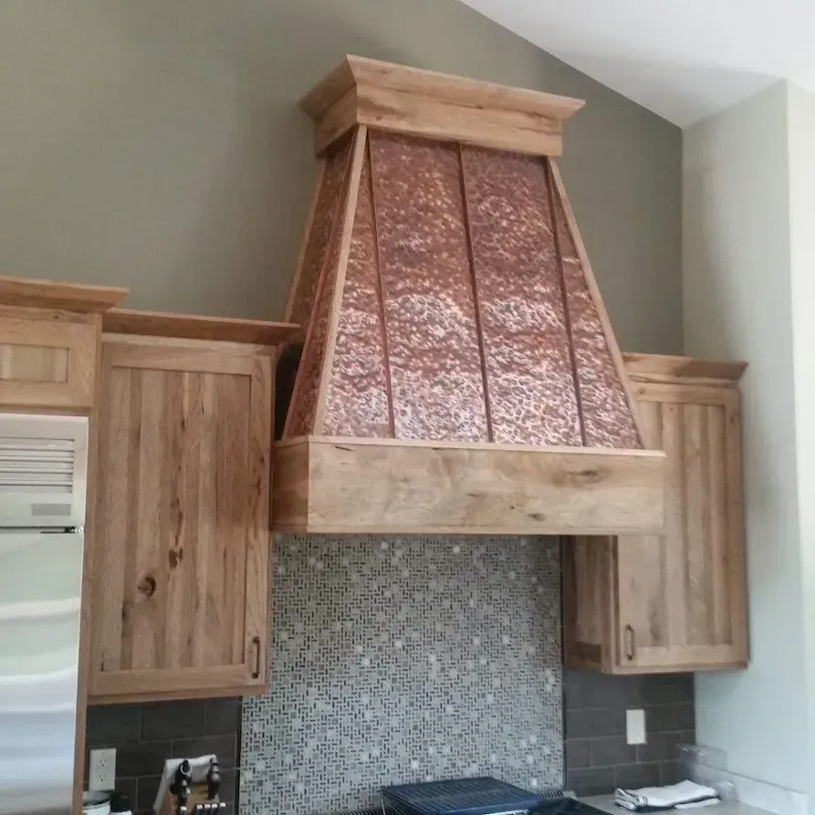 Copper range hood with wood trim in a kitchen. Custom copper range hood between wooden kitchen cabinets over a tiled backsplash.