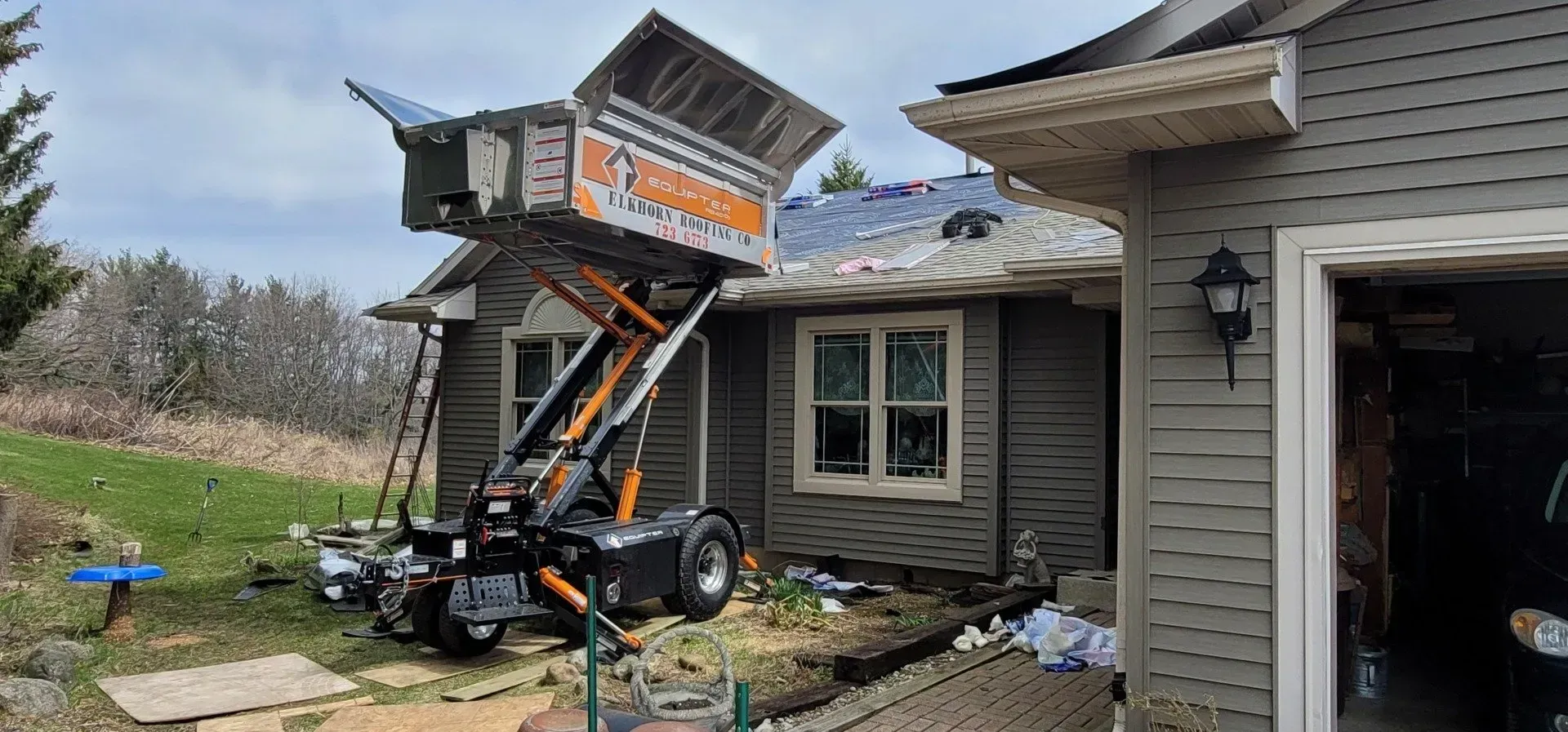 A roofing tear-off project. A debris lift is used to remove old roofing from a house. Dumpster lifted by excavator beside a house, with an open garage and roof work in progress