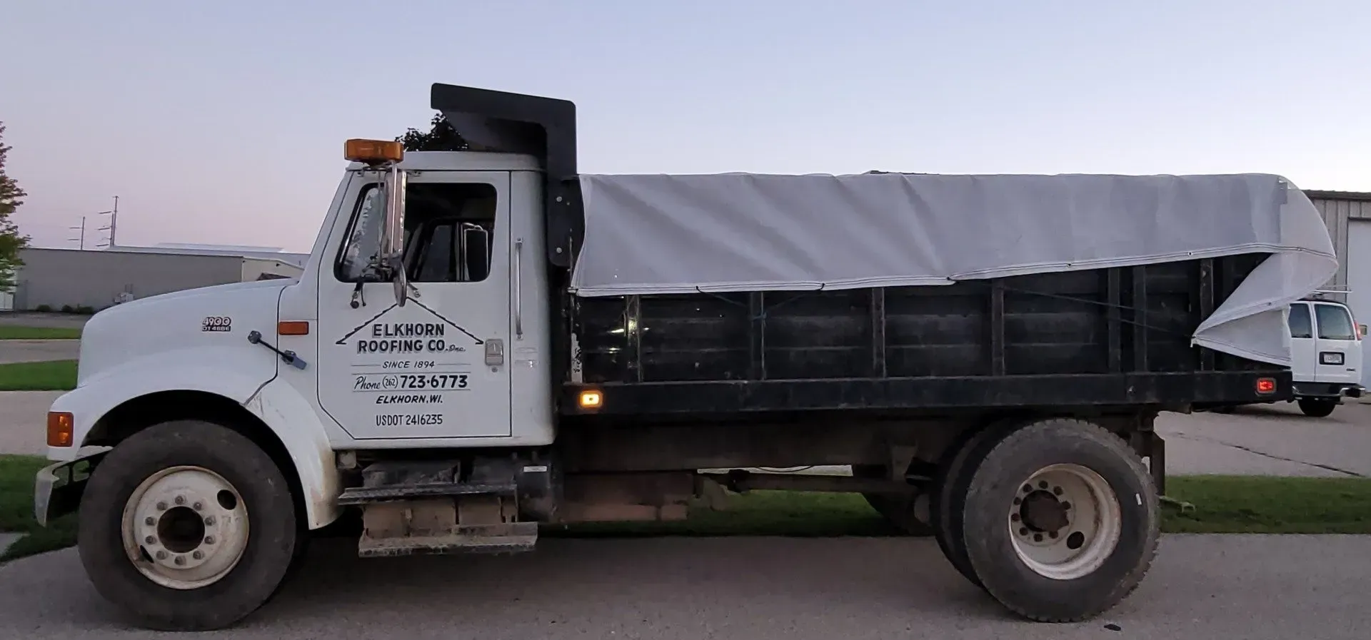 White dump truck with black bed and tarp on side of road. White dump truck with black bed and gray tarp parked on a road at dusk.