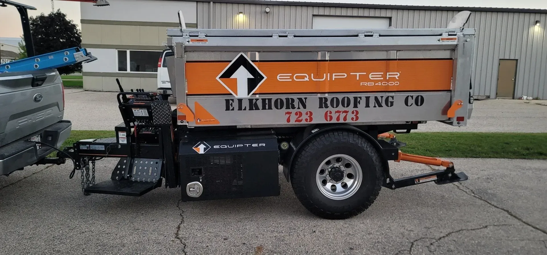 Truck towing a roofing trailer with Elkhorn Roofing Co. logo. Orange roofing truck with equipment trailer parked on a street, labeled Elkhorn Roofing Co.