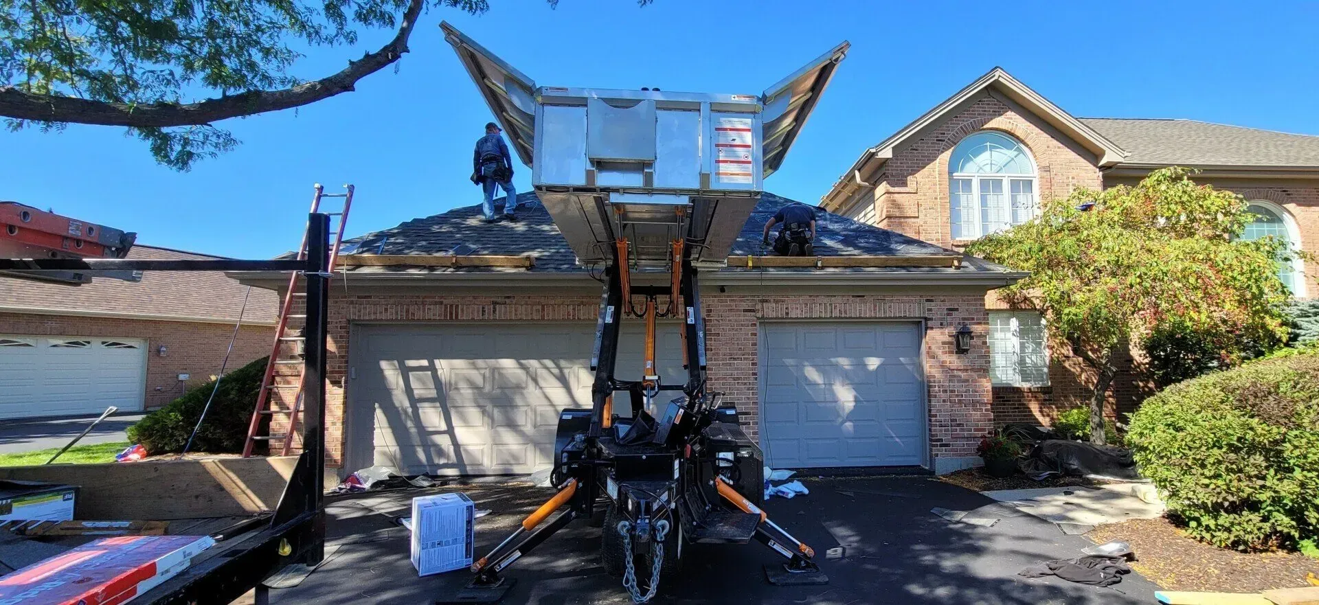 A roofer on a house roof is working with a lift. Sunlight and blue sky are in the background. Damaged roof over a suburban two-car garage with debris and a trailer in the driveway