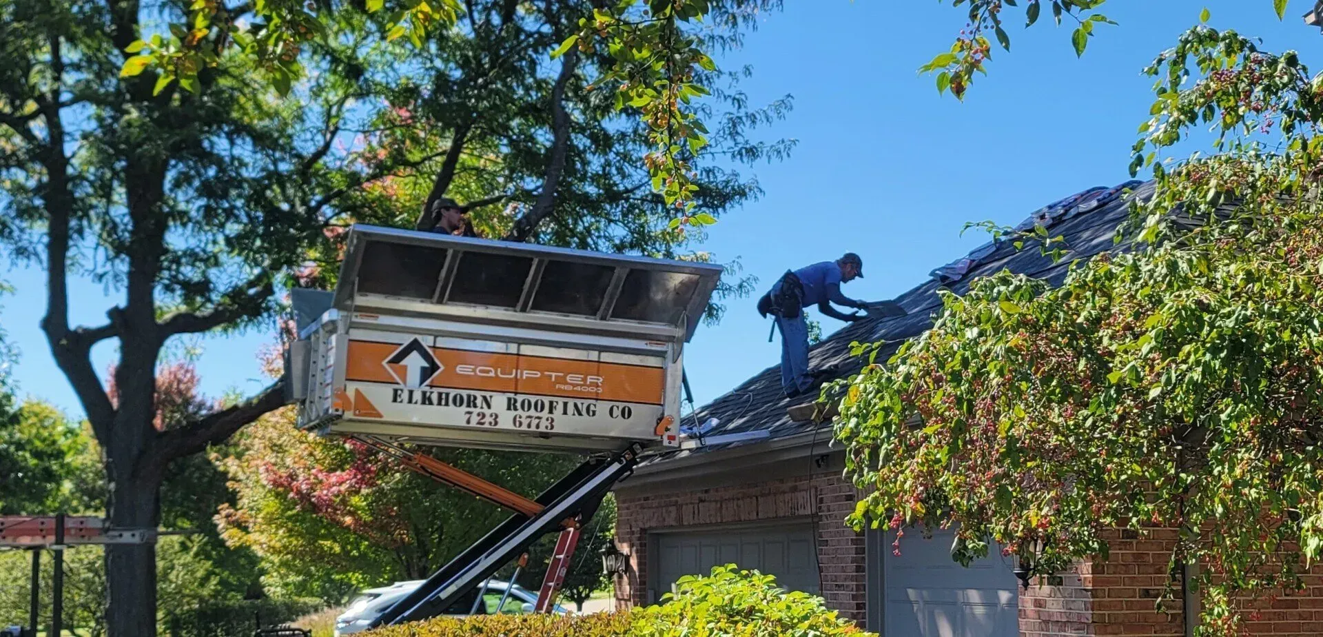 A roofer on a house roof is working near a dumpster being hoisted by a lift, sunny day. Worker on a roof installing a sign above a brick building, with trees and blue sky in the background