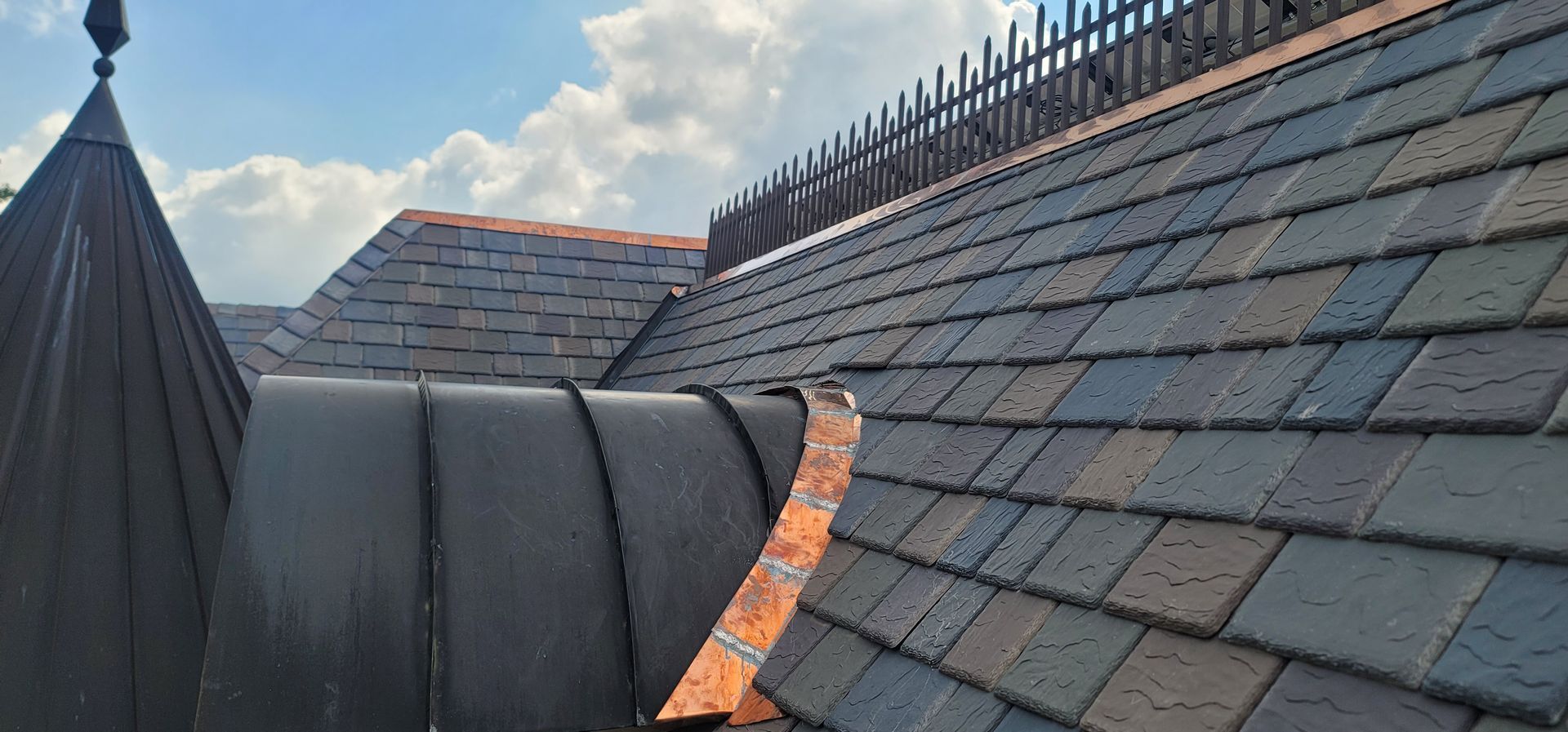 Rooftop with dark slate tiles, a curved copper seam, and a pointed black turret against the sky