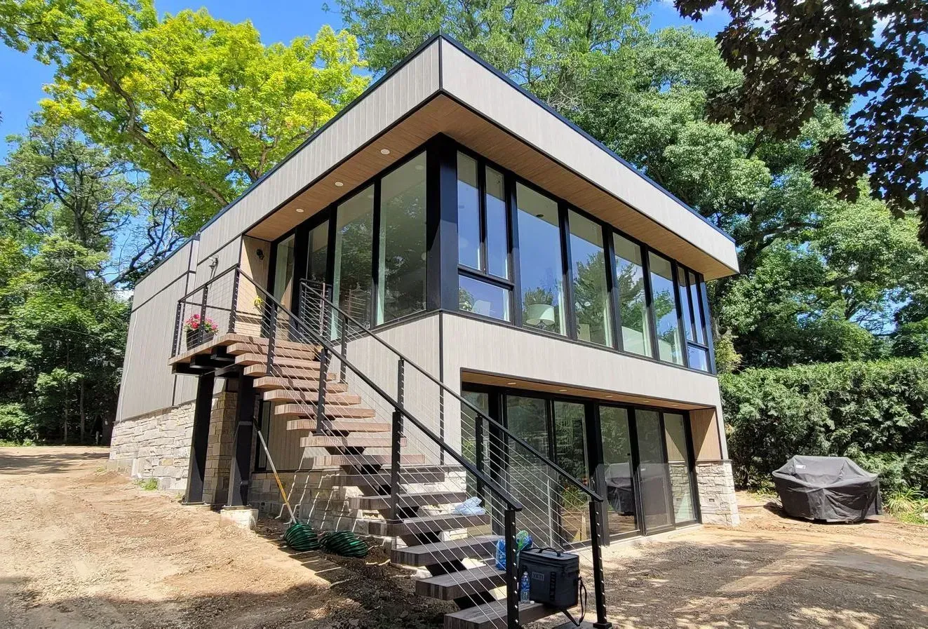 Modern two-story house with glass walls, exterior stairs, and surrounded by trees. Modern two-story house with large windows, exterior stairs, and a wooded hillside setting