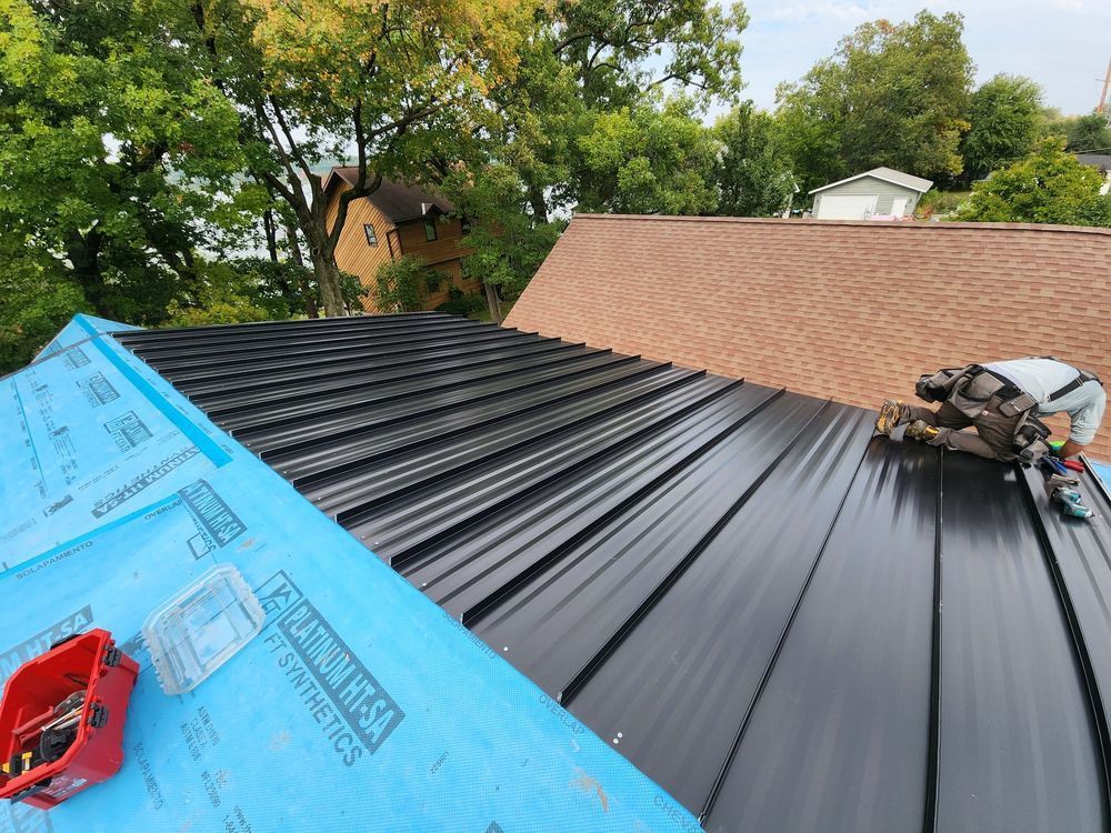 Roofing work on a house, with black metal panels being installed beside blue underlayment and tools.
