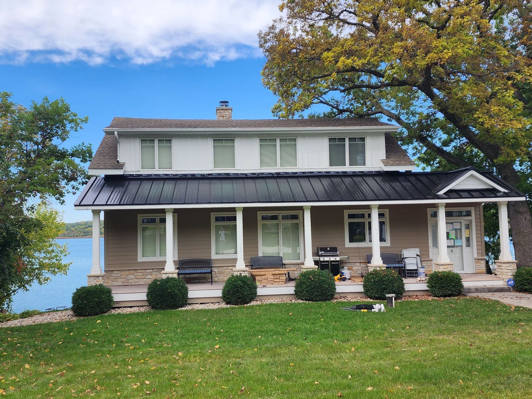 White two-story house with porch and dark roof on a grassy hill by the water under a blue sky