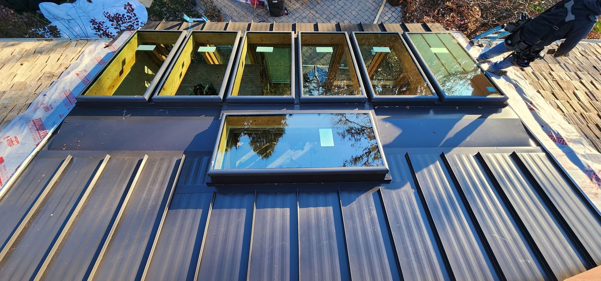 Tilted rooftop with metal panels and a row of skylight windows under blue sky.