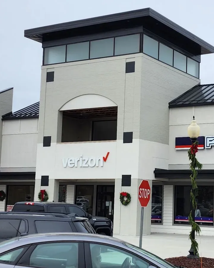 Verizon store exterior with gray brick facade, open second story, and black roof. A stop sign and garland visible. Verizon storefront in a shopping center, with parked cars and a stop sign in front