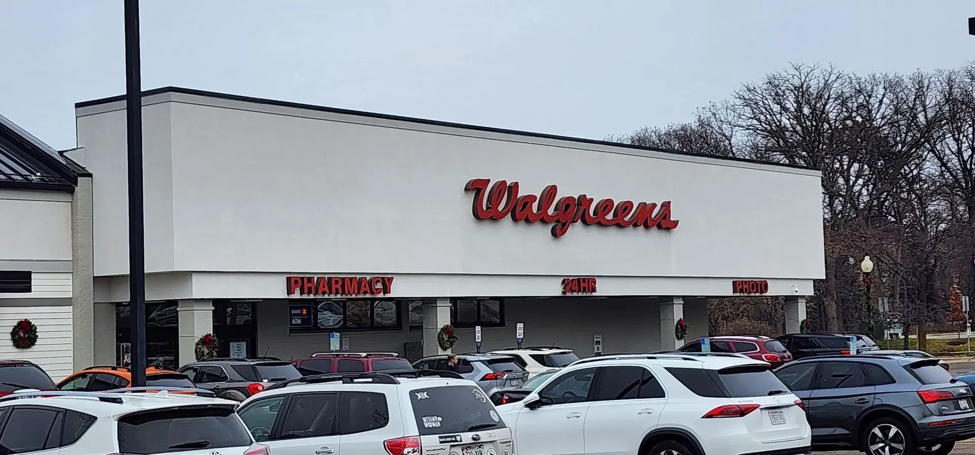 Exterior of a Walgreens store with the red logo on a white facade. The pharmacy, and shop is visible, and the parking lot has vehicles. Walgreens storefront with cars parked outside on a snowy day