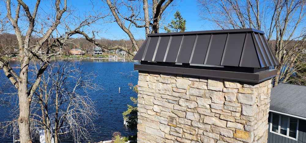 Stone chimney by a blue lake with leafless trees and a dark metal cap under a clear sky