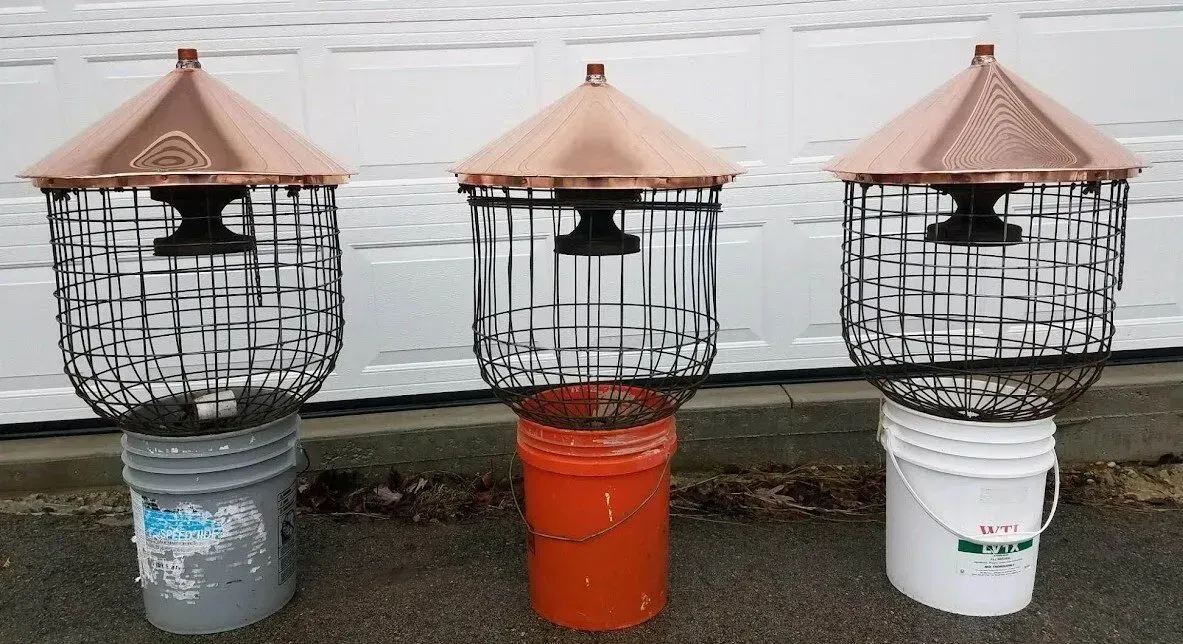 Three copper-topped bird feeders with wire cages sit on buckets, next to a white surface. Three wire bird feeders with copper roofs sit on colored buckets against a wall.