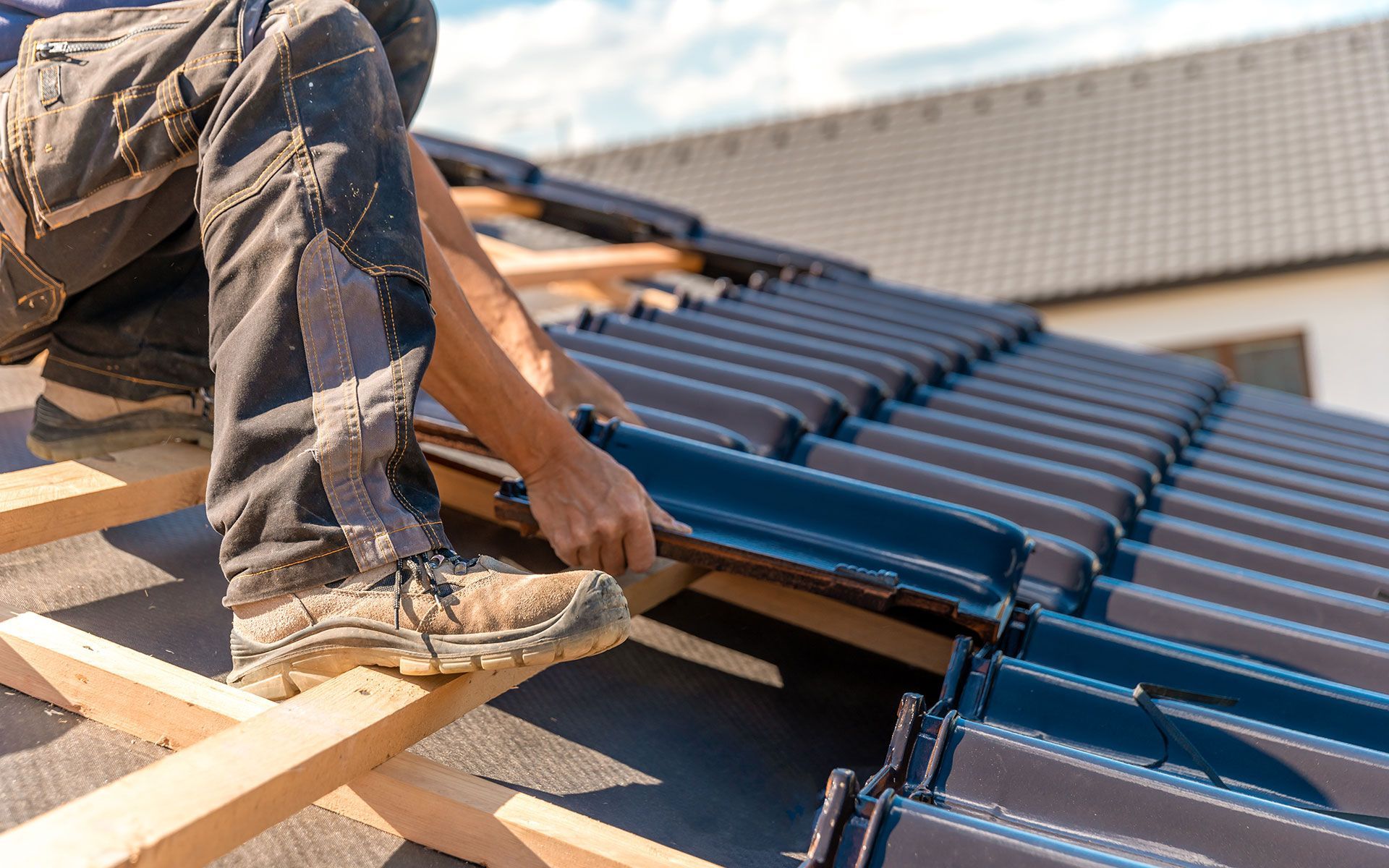 Worker installing dark blue roof tiles on a roof, kneeling on wooden supports