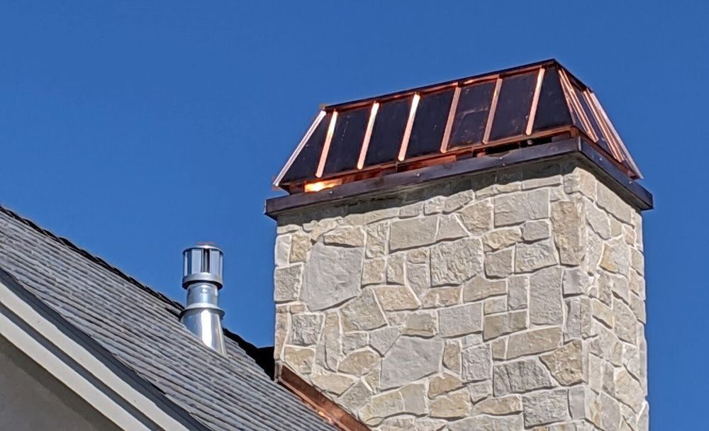 Stone chimney with copper cap beside a gray roof against a blue sky