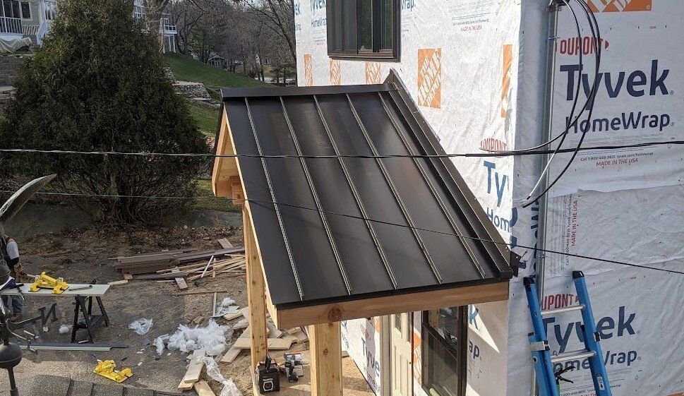 House under construction with dark metal roof panels and Tyvek house wrap, surrounded by debris and tools.