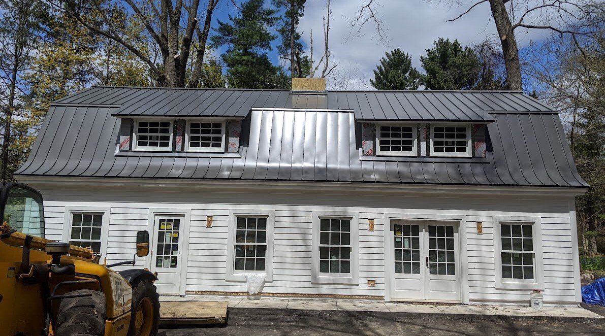 White house with dark gray metal roof and dormer windows, seen from the front in a wooded yard.