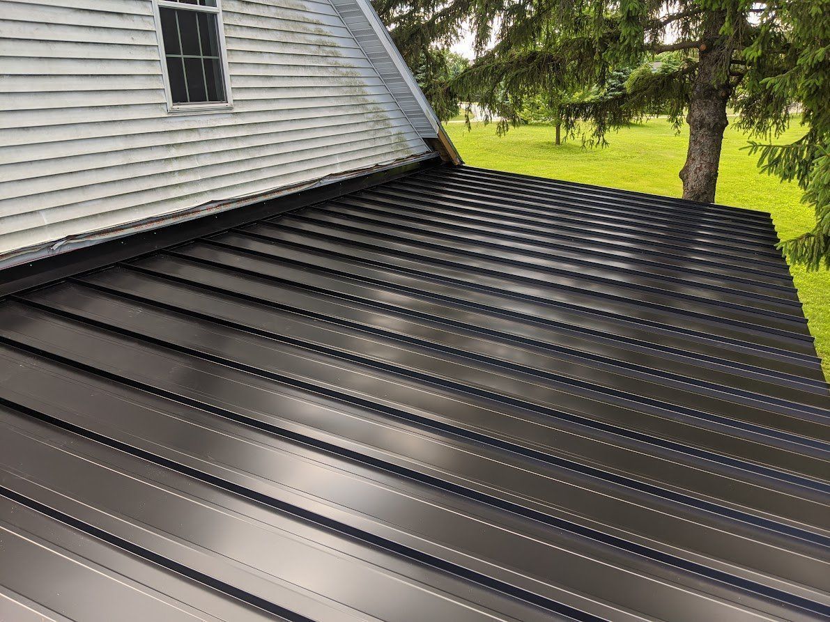 Black metal roof on a house, contrasting with the gray siding and green trees in the background. Black metal roof extension on a gray house beside a green lawn and trees