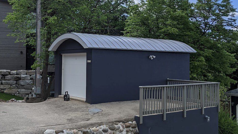 Small blue shed with curved metal roof beside a wooden deck and trees outdoors