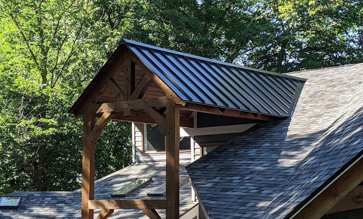 Wooden porch roof with dark blue metal roofing on a house, surrounded by leafy trees.