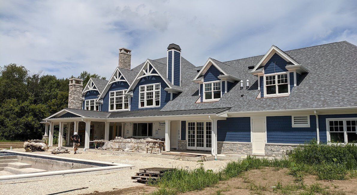 Large gray house under construction with dormer windows, stone chimney, and unfinished front yard